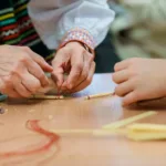 Zbliżenie na dłonie twórczyni ludowej. Kobieta pomaga dziecku wiązać słomę.