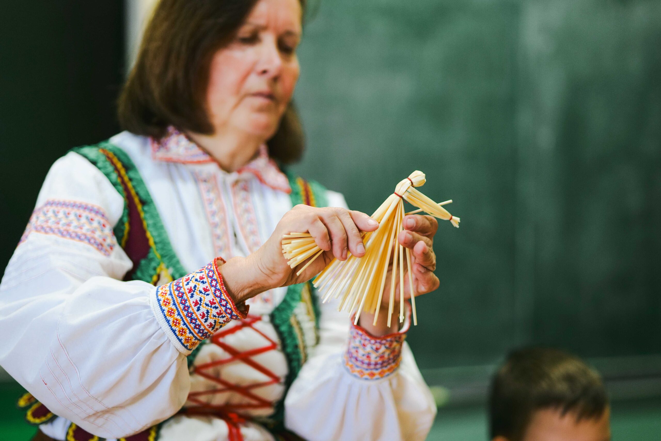 Twórczyni w stroju ludowym z haftowanymi mankietami trzyma w ręku ozdobę ze słomy - małą babę jagę na miotle.